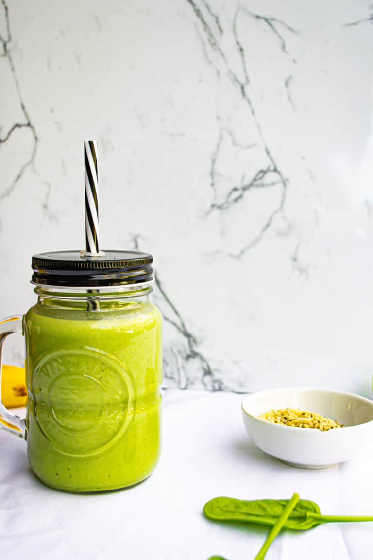 Creamy green smoothie in mason jar with striped straw, served with bowl of granola, on white table with marble wall background, highlighting healthy breakfast recipes and nutritious smoothies.