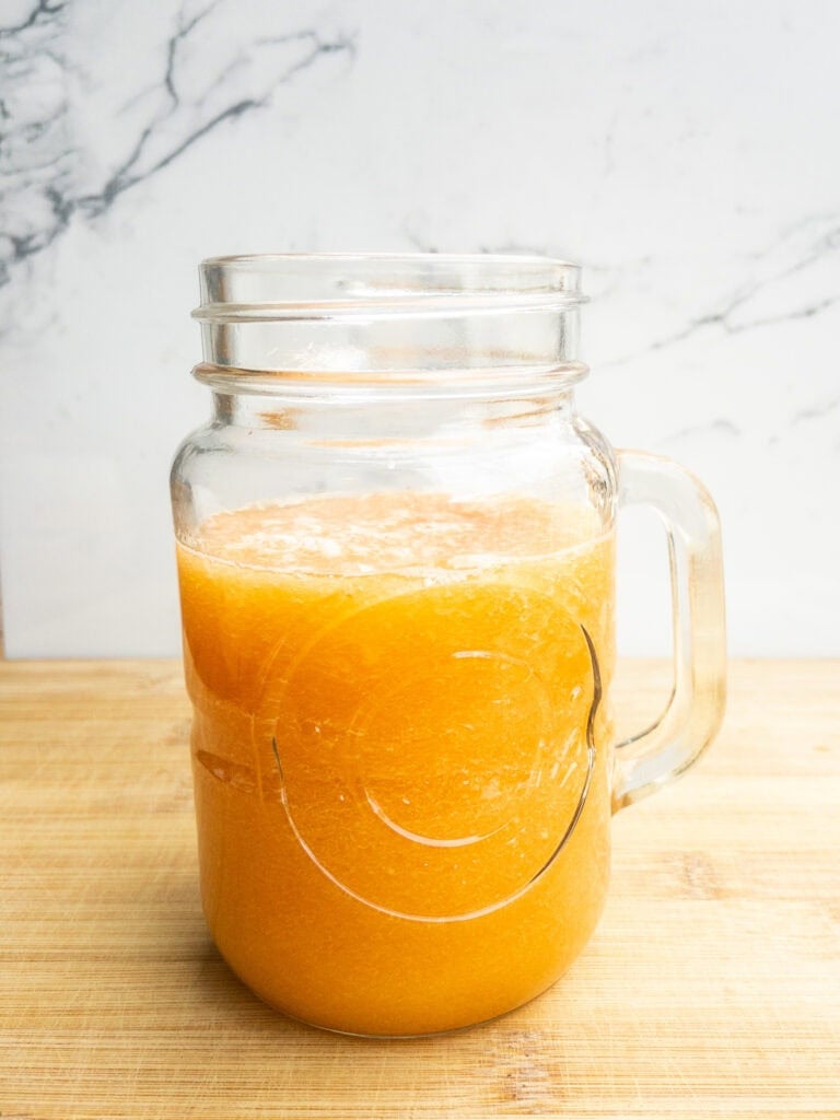 Fresh orange juice in a glass mason jar with a handle, showcasing vibrant color and pulp, on a wooden surface with a white marble background.