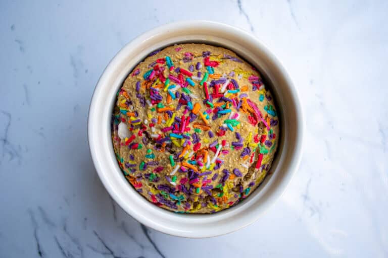 Colorful sprinkled birthday cake in a white bowl on a marble surface.