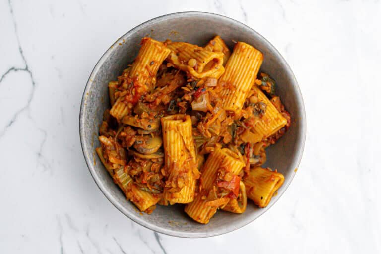 Creamy vegetable pasta in a gray bowl, topped with rich tomato sauce, cooked with zucchini, mushrooms, and onions, served on a white marble surface.