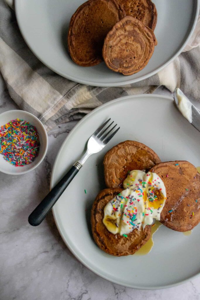 Top view of healthy chocolate pancakes on a plate with yogurt, sprinkles, and syrup as toppings.