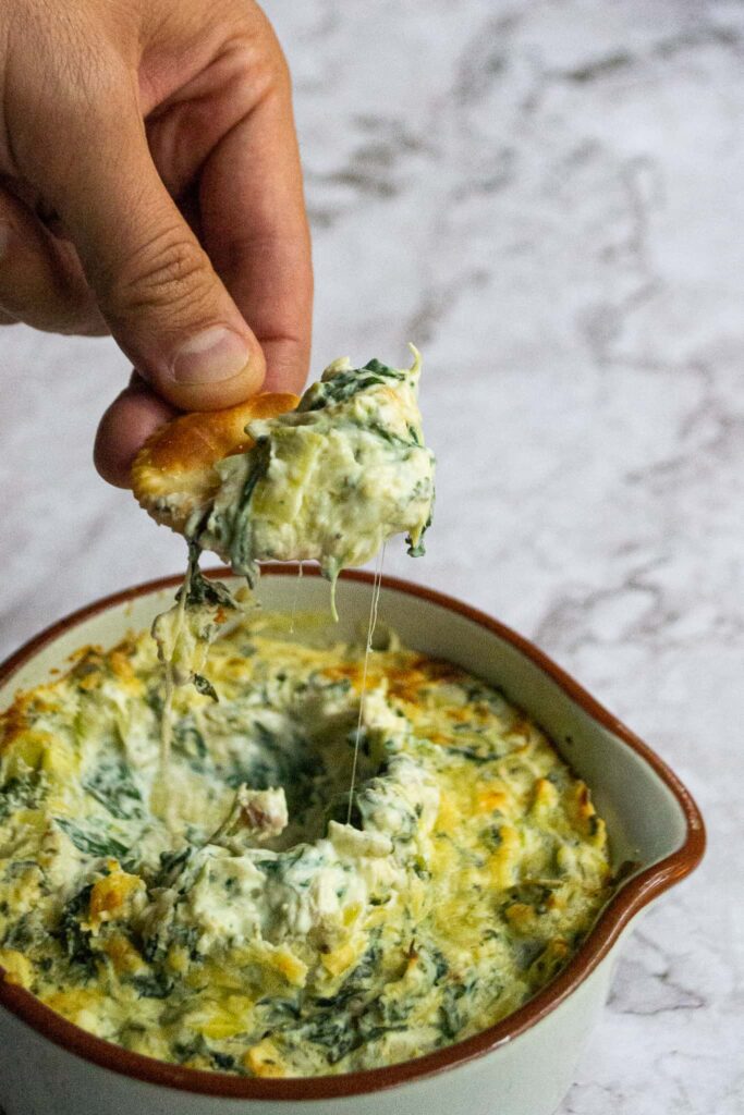 Healthy spinach artichoke dip in a bowl with a person scooping some onto a cracker.