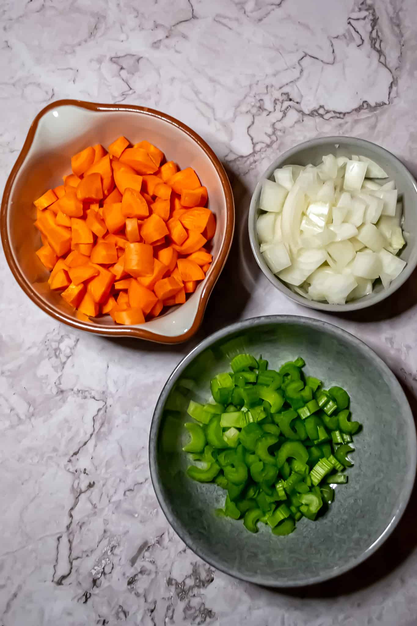 Top view of a bowl of carrots, a bowl of diced onions, and a bowl of sliced celery