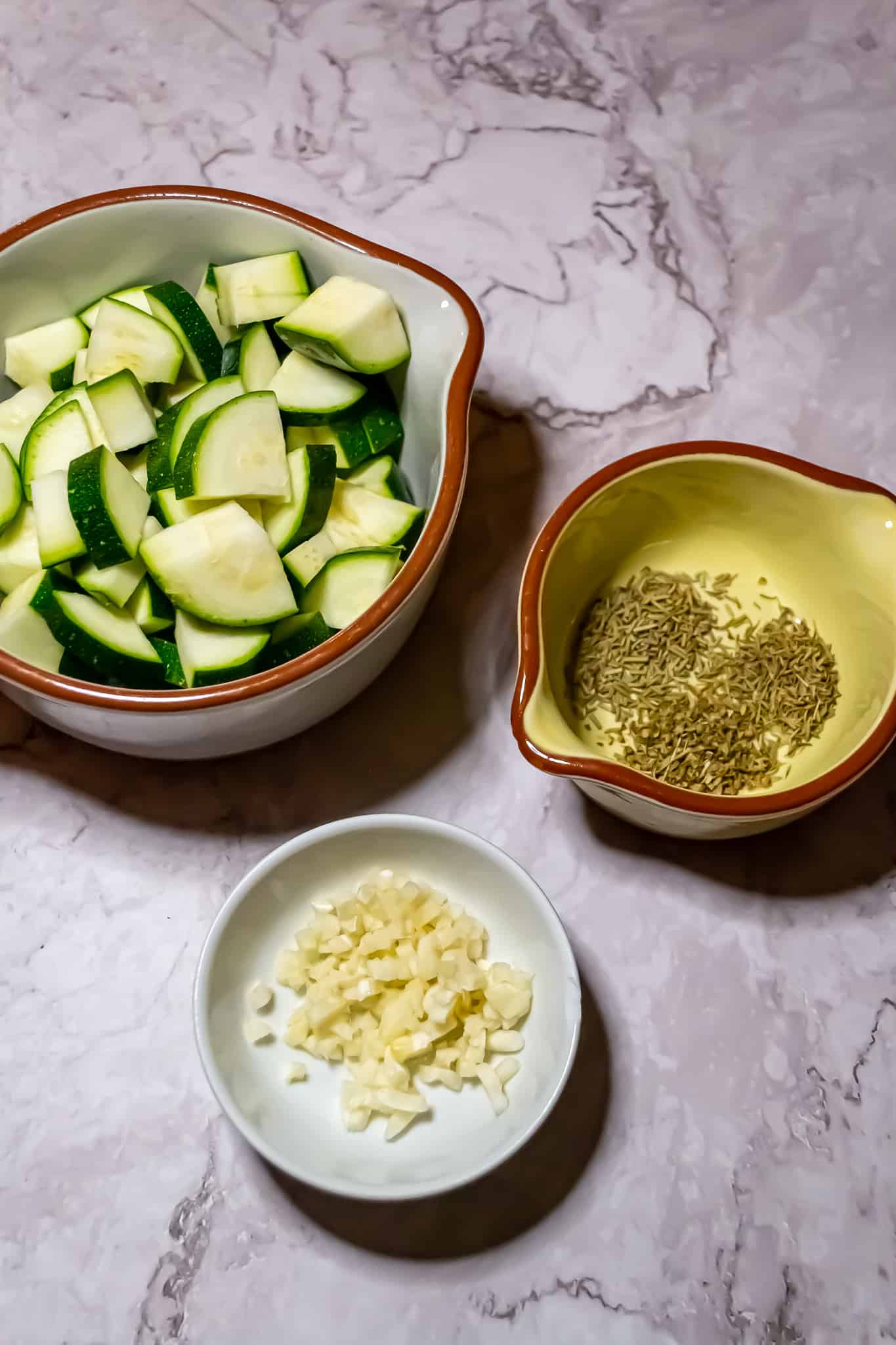 Top view of a bowl of zucchini slices, a small bowl of diced garlic, and a bowl of herbs.