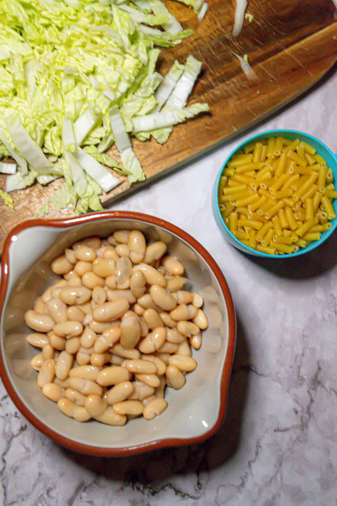 Top view of a bowl of cannellini beans, napa cabbage on a cutting board, and a measuring cup of macaroni.
