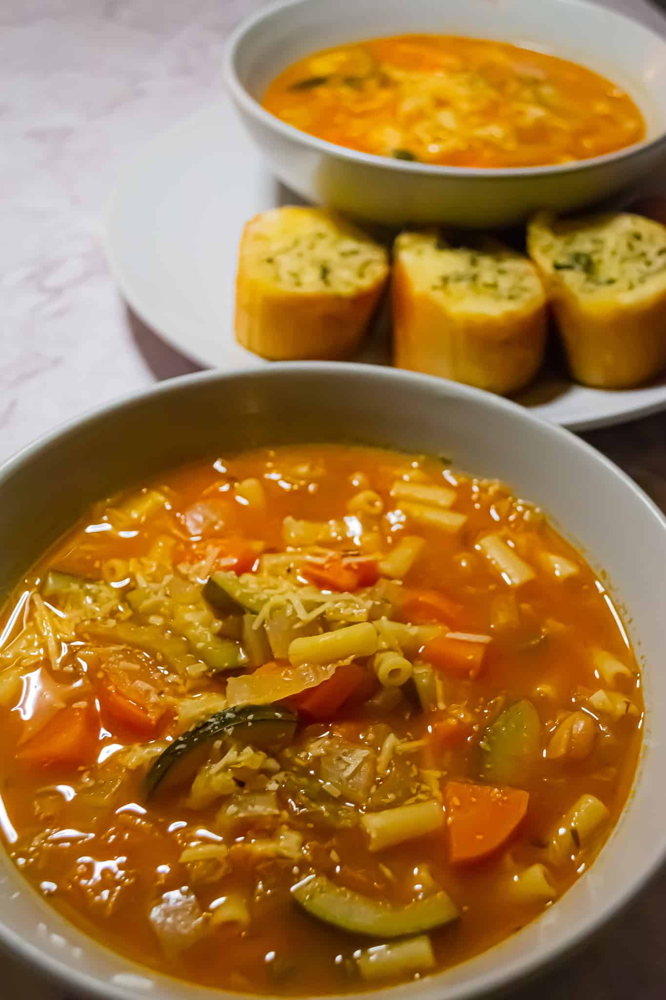 Close up shot of a bowl of healthy minestrone soup. In the background, you can see another bowl of soup on a plate with a side of garlic bread.