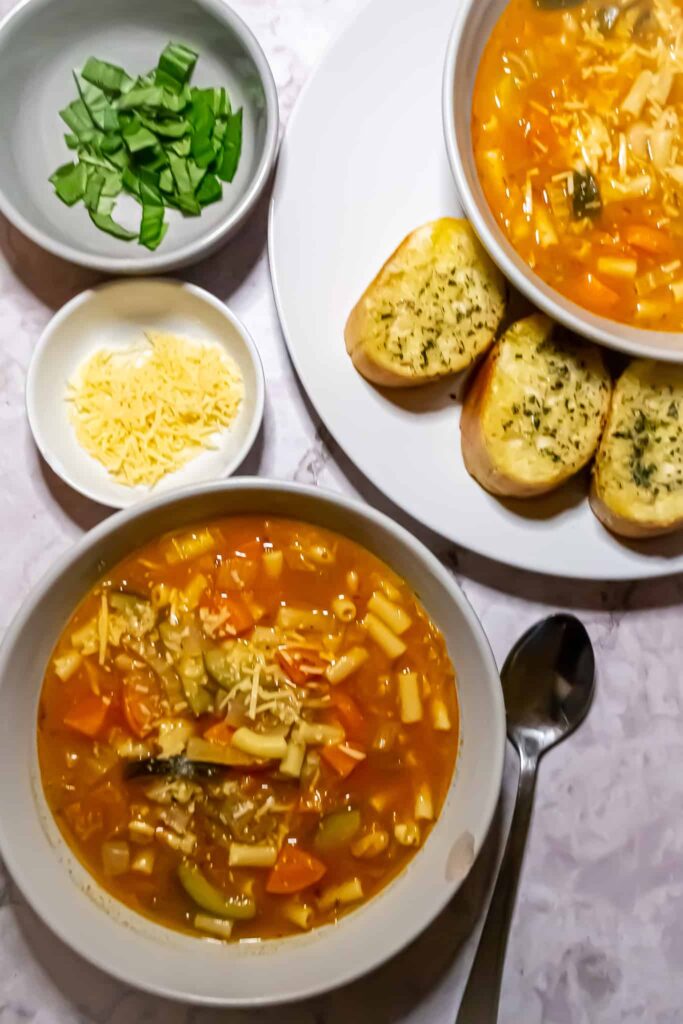 Two bowls of healthy minestrone soup on a marble table. There is a small bowl of basil, a small bowl of parmesan, and a plate of garlic bread next to the bowls of soup.