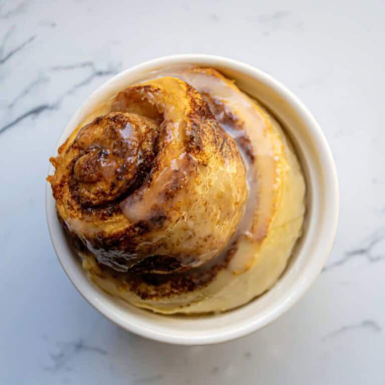 Cinnamon roll ice cream topped with cinnamon, sugar, and caramel swirls in a white bowl on a marble surface, perfect for dessert or sweet treat.