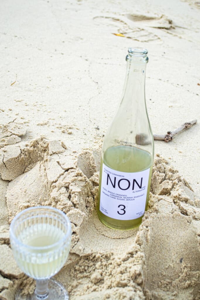 A bottle of NON 3 sitting in the sand besides a full glass of it.