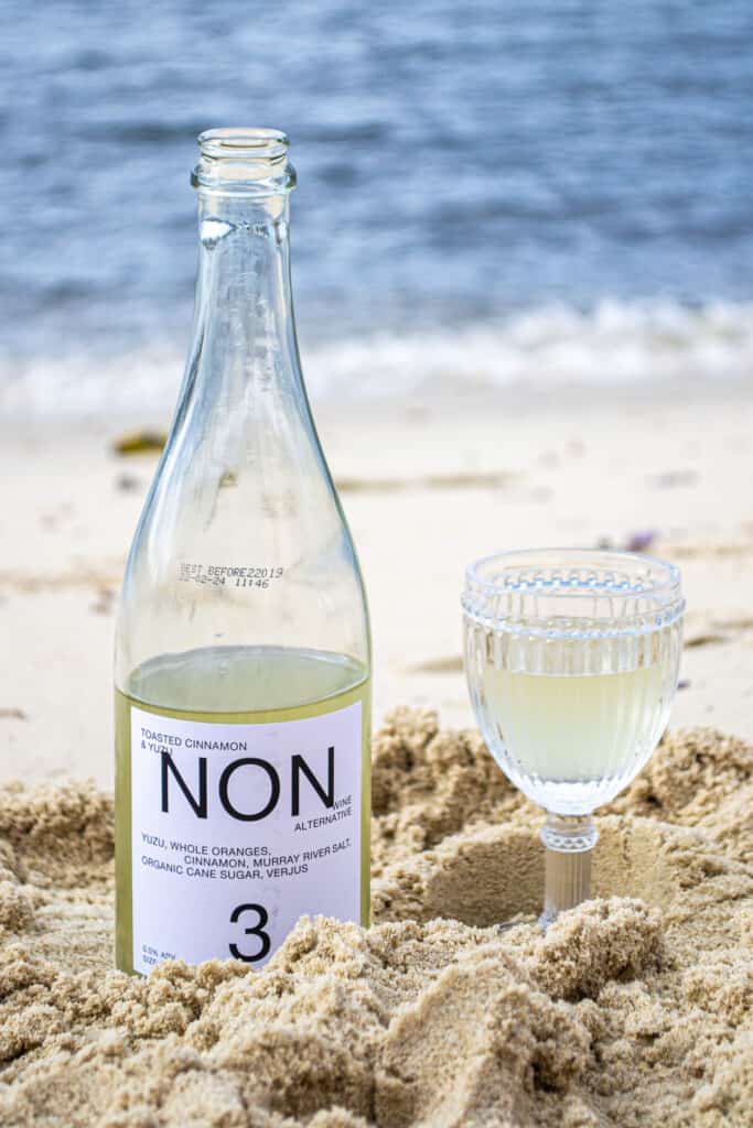 A bottle of NON 3 sitting in the sand next to a glass of NON 3, with the ocean in the background.