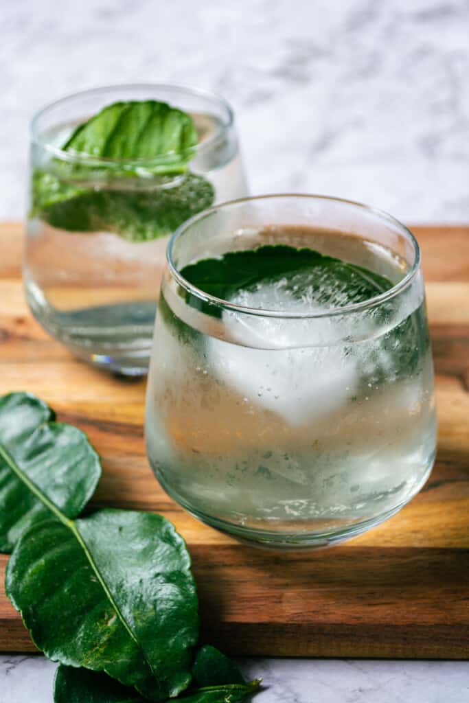 A front shot of two yuzu mocktails in glasses, sitting on a wooden board on top of marble with a kaffir lime leaf in the foreground.