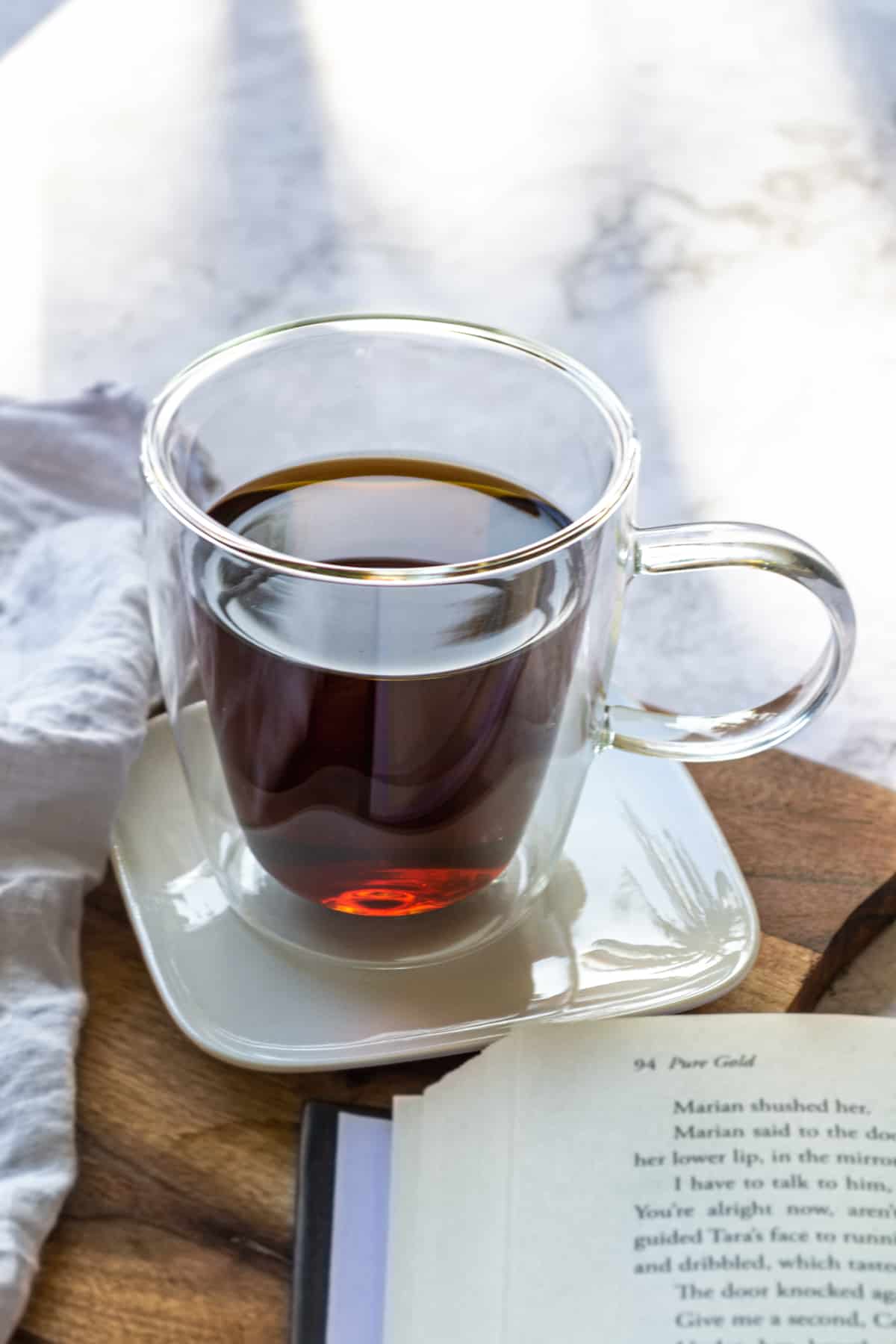 A glass mug of dandelion tea sitting on a white square plate with an open book in the foreground.