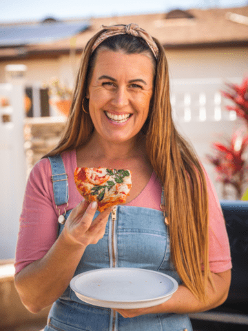 Photo of chef Sarah Glover in a pink shirt and overalls eating a slice of her cauliflower pizza.