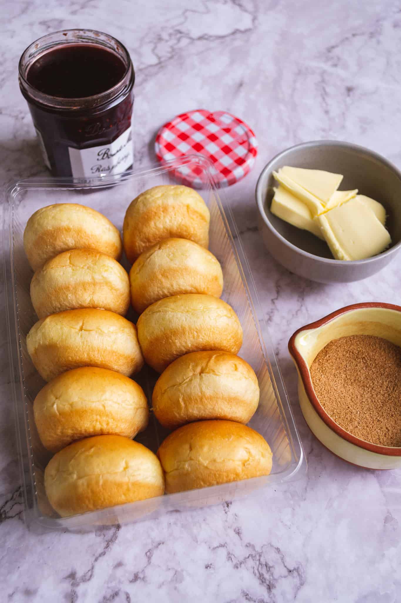 Overhead shot of the ingredients to make semi homemade brioche donuts on a marble table, including jam, brioche, butter and cinnamon sugar.