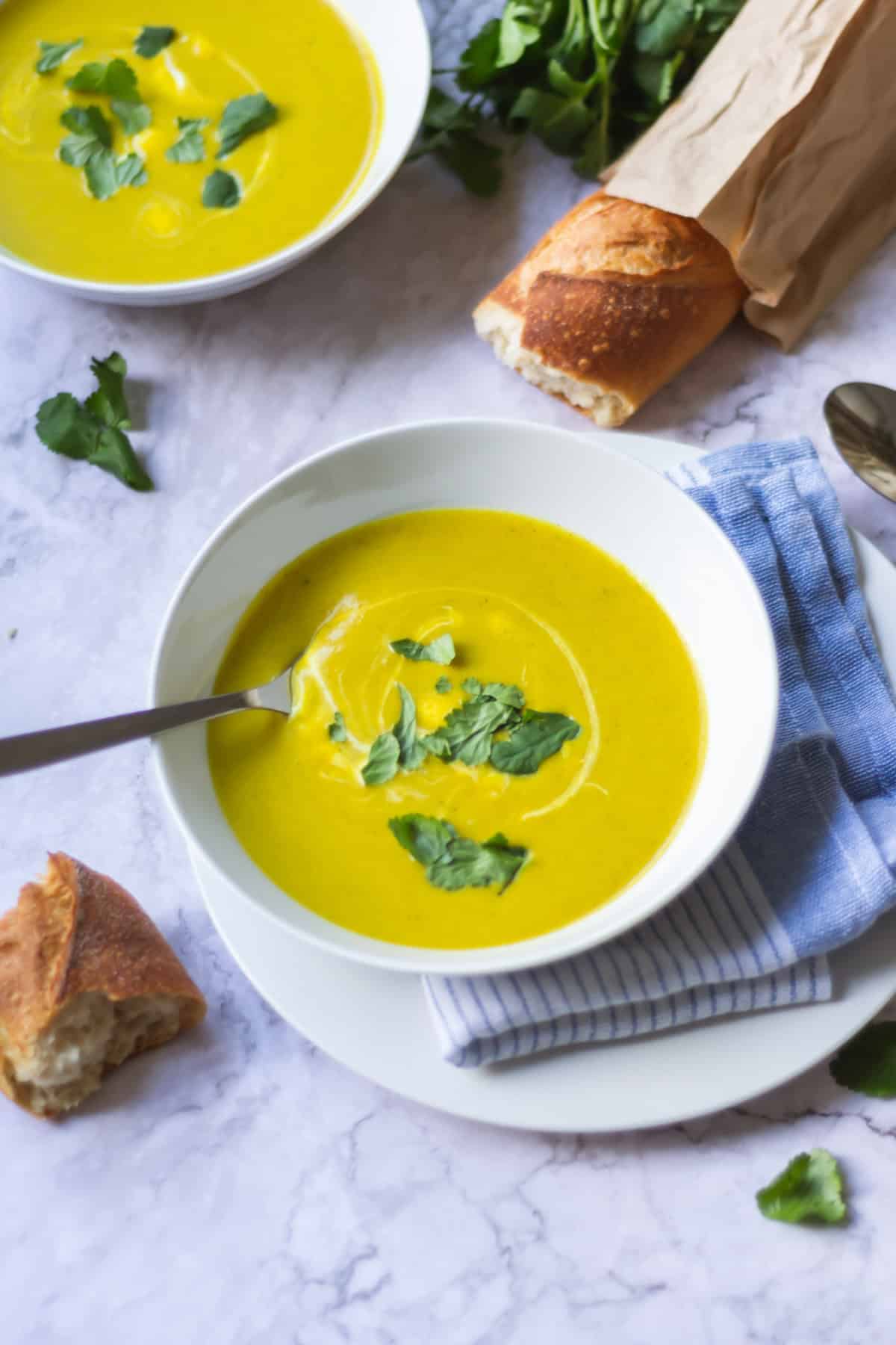 An overhead view of two bowls of butternut squash and broccoli soup on a marble table with a baguette and coriander.