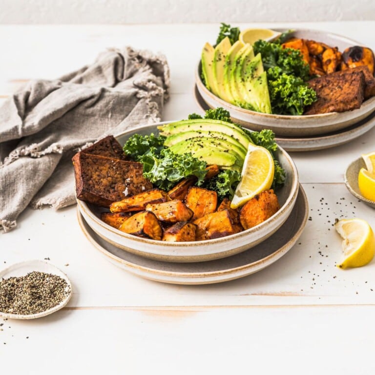 Savory roasted sweet potatoes, avocado slices, kale, and lemon wedges in ceramic bowls, prepared for a healthy meal or meal prep, featured on a white rustic table.