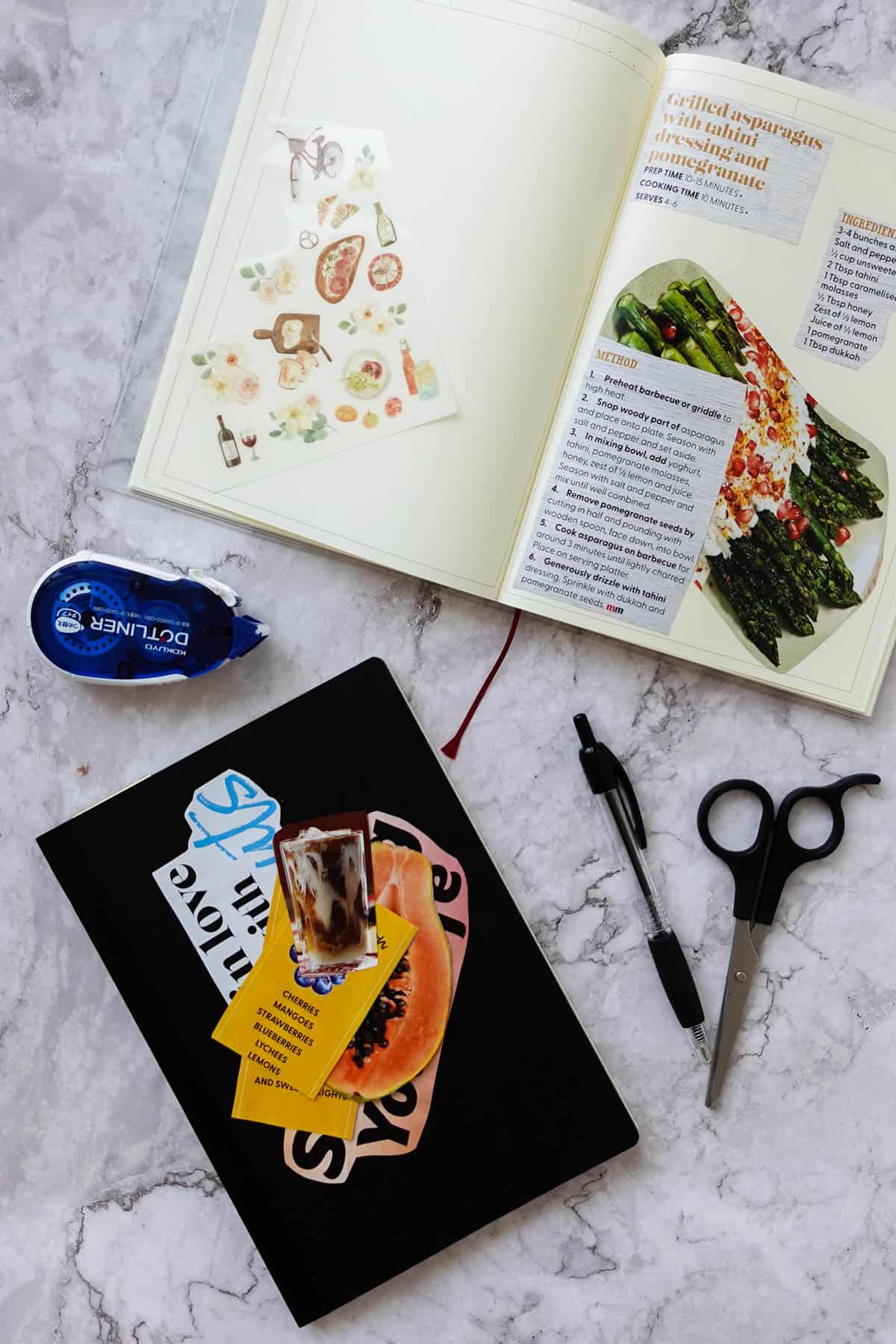 Fresh vegetables and recipe book for grilled asparagus with tahini dressing on a white marble surface, featuring scissors, pen, and a foodie magazine for healthy meal ideas.