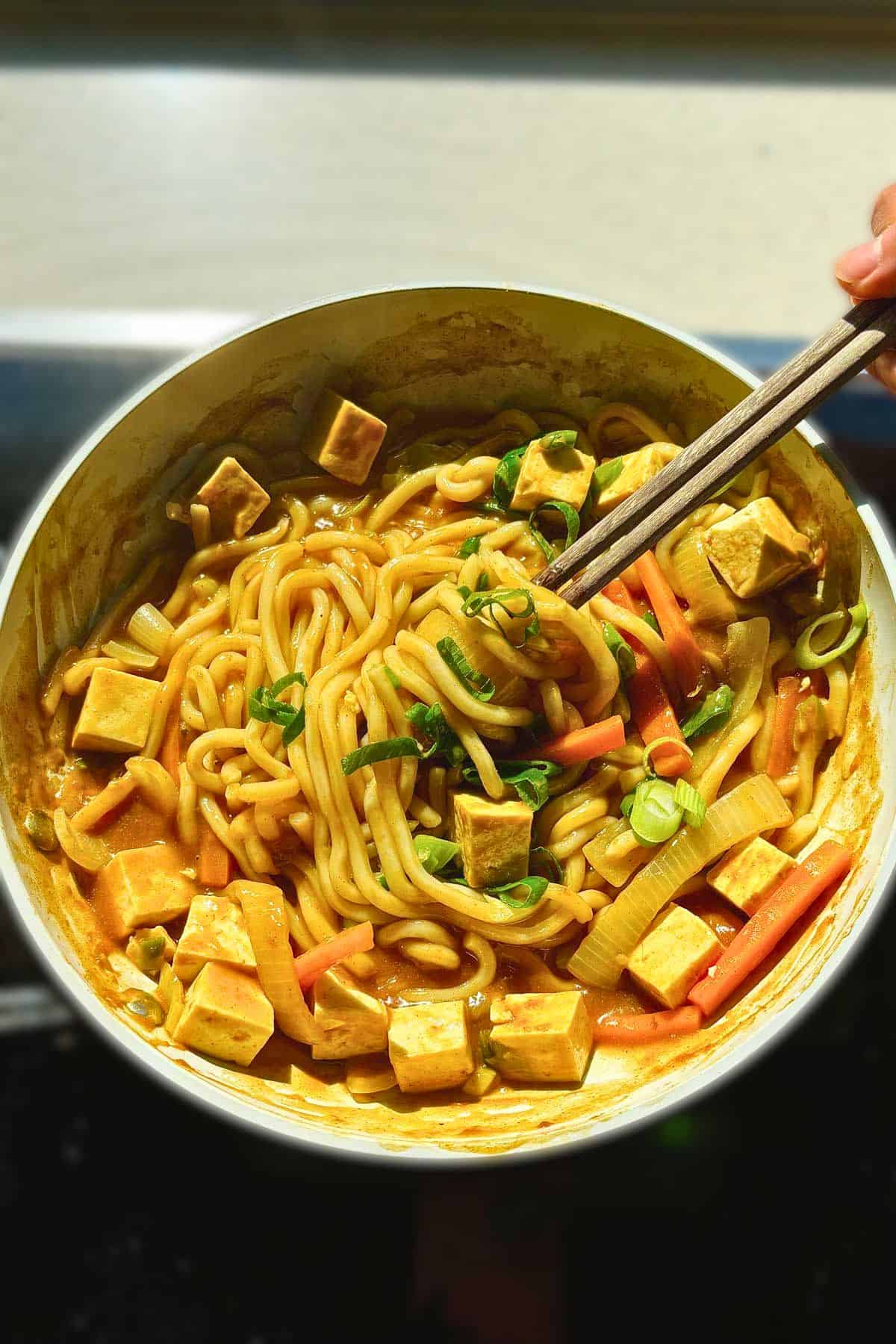 Overhead shot chopsticks taking a bite of stir-fried curry udon from a pan with tofu, carrots, and scallion.