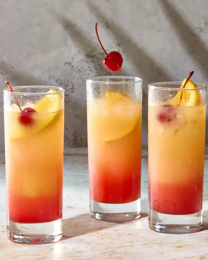Three glasses of Tequila Sunrise served in highball glasses, with orange wedges and cherries.